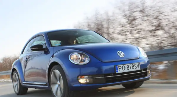 Niebieski, nowy Beetle pędzi po drodze, za nim rozmyty krajobraz drzew.