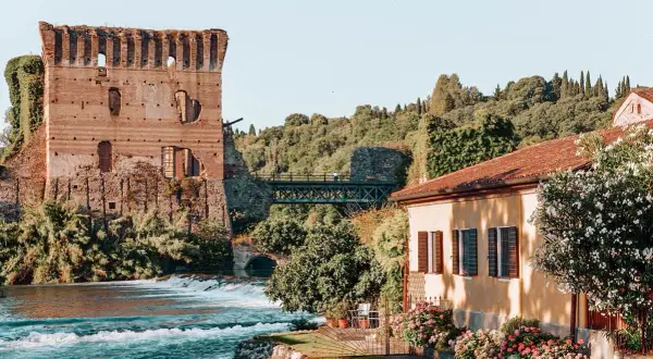 Rovine di un castello medievale e una casa con fiori colorati, un fiume e un ponte: un quadro perfetto dei borghi veneti da visitare.