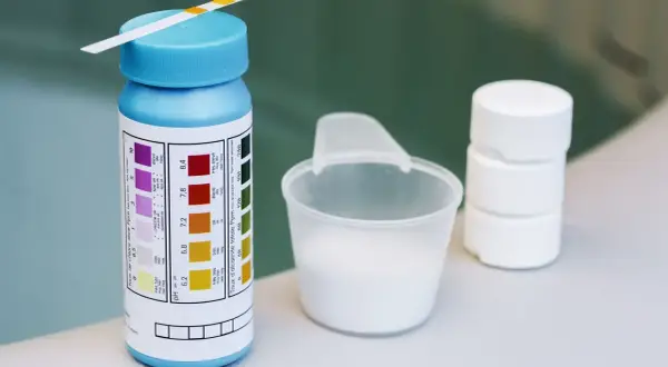 Teststreifen für den ph-wert pool, daneben ein Messbecher mit weißem Pulver und Tabletten.