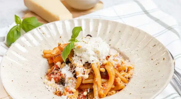 En tallrik med pasta alla norma, toppad med ricotta och basilika. Bredvid syns en bit parmesanost och en skål med pinjenötter.