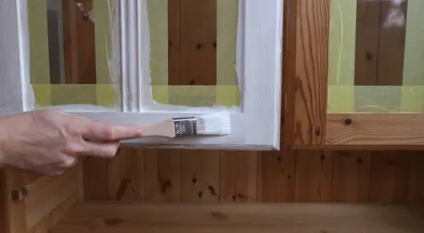 En hand målar en vit kant på en trämöbel. Maskeringstejp skyddar träet.