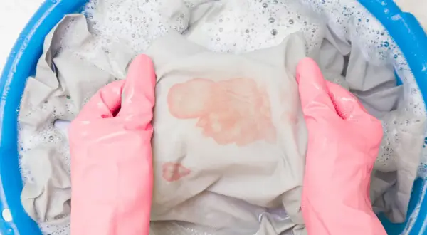 Mani con guanti rosa che lavano un vestito macchiato di rosso in una bacinella blu con acqua saponata. Ecco come sbiancare i vestiti che hanno preso colore.