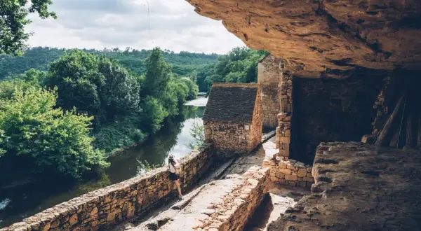 Une femme explore un village troglodytique en Dordogne, au bord d'une rivière paisible. Une belle idée de que faire en Dordogne.