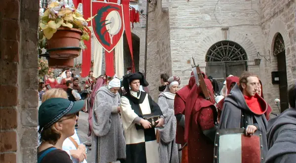 Persone in costume medievale sfilano in una strada acciottolata, con stendardi rossi e bianchi che sventolano. La scena evoca il **calendimaggio significato**, una festa di primavera.