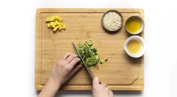 Hände schneiden Brokkoli für Babybrei. Daneben liegen Kartoffelwürfel, Haferflocken und Öl.