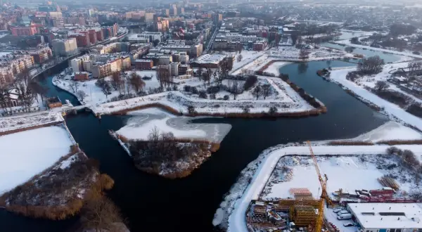 Zimowy krajobraz dolnego miasta Gdańsk z rzeką, ośnieżonymi budynkami i placem budowy z dźwigami.