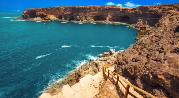 Fuerteventura na co uważać? Klify i turkusowe morze, ścieżka z drewnianą balustradą prowadzi w dół. Uważaj na nierówny teren.