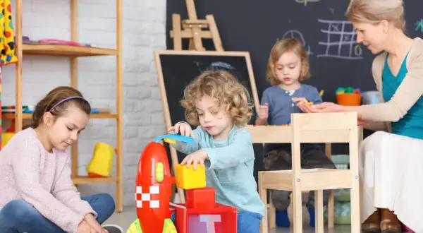 Dzieci bawią się klockami i rakietą. Czy praca w żłobku jest ciężka? Widok radosnych maluchów i opiekunki sugeruje, że niekoniecznie.