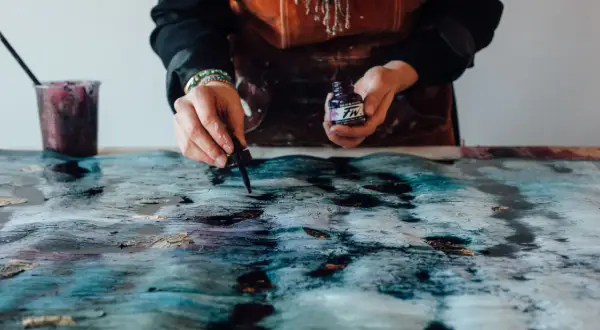 Mani che creano un'opera d'arte con colori acrilici su plastica, gocciolando inchiostro viola su una superficie texturizzata blu e oro.