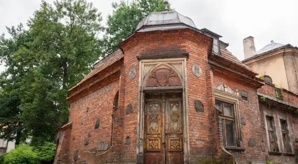 Stary ceglany budynek z ozdobnymi drzwiami i medalionami, otoczony zielenią.
