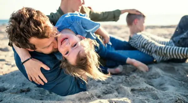 Rodzina na plaży, ojciec tuli córkę, która śmieje się do aparatu. W tle mama i syn. Idealne miejsce na jastrzębia góra atrakcje dla dzieci.
