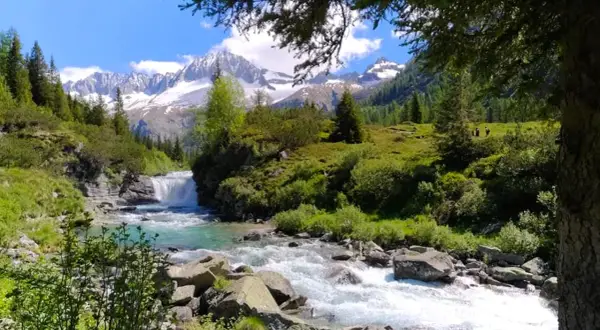 Un torrente impetuoso scorre tra rocce e vegetazione lussureggiante, con una cascata sullo sfondo. Le cime innevate delle montagne dominano il paesaggio del Val di Fumo, suggerendo l'alta altitudine.