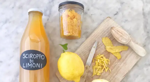 Bottiglia di sciroppo di limoni, limoni freschi, scorze e un coltello su tagliere. La sciroppo di limone ricetta della nonna è pronta!