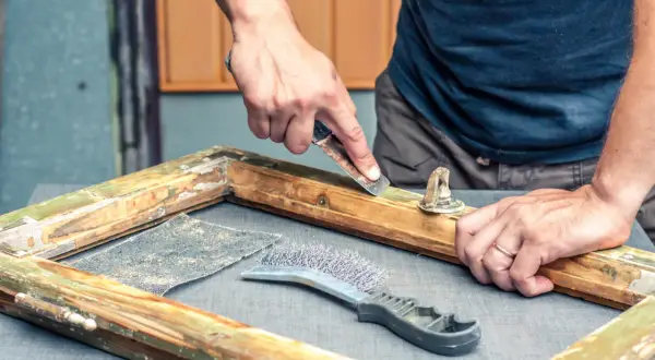 Mani esperte raschiano un vecchio telaio di legno per riportare il legno al naturale, usando carta vetrata e spazzola metallica.