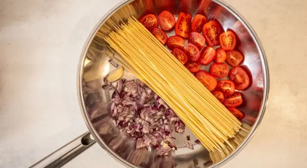 Zutaten für ein One-Pot-Gericht: Spaghetti, gehackte rote Zwiebeln, Kirschtomaten und eine Knoblauchzehe in einer Pfanne.