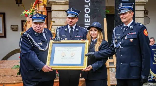 Uroczystość wręczenia nagród. Strażacy OSP w mundurach, z medalami, odbierają dyplom. Widać, jak nosić medale na mundurze OSP.