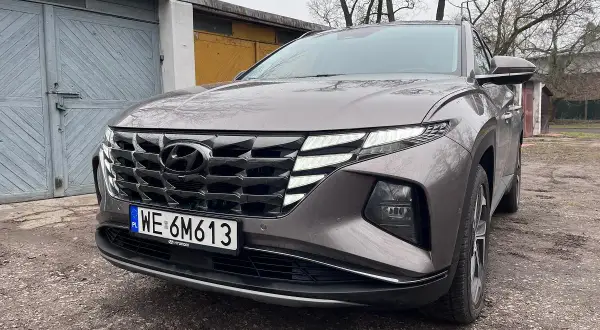Szary Hyundai Tucson Hybrid z charakterystycznym grillem i światłami LED stoi przed garażami.