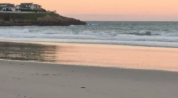 Ocaso en la playa areoura. Olas rompen suavemente en la arena dorada, reflejando el cielo rosado. Casas blancas coronan el acantilado.