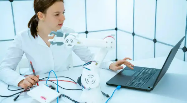 Junge Frau mit einem Roboterarm und Laptop, die an einem Projekt für ihr Robotik Studium arbeitet.