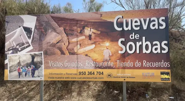 Cartel anunciando las Cuevas de Sorbas, con imágenes de exploradores y formaciones rocosas. Ofrecen visitas guiadas, restaurante y tienda.