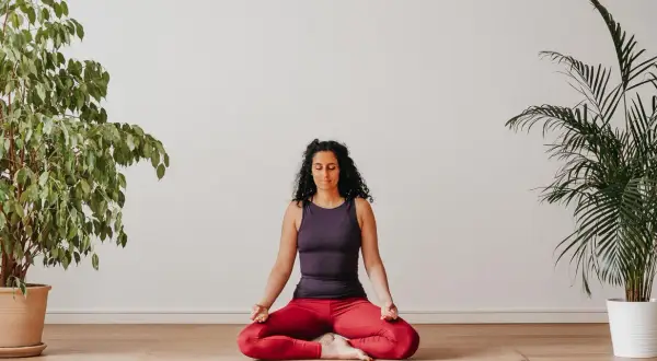 Frau praktiziert Yoga zu Hause im Lotussitz, umgeben von Pflanzen.