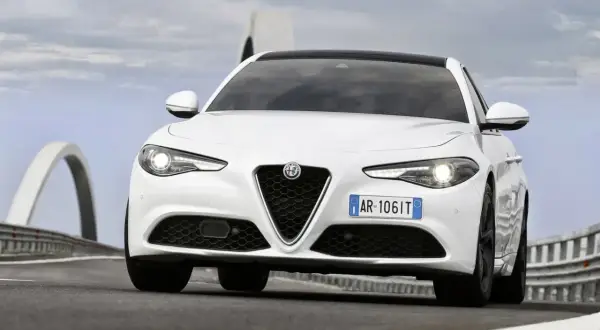 Biała Alfa Romeo Giulia Quadrifoglio z czarnym dachem pędzi po drodze z mostami w tle.
