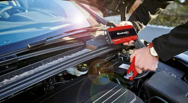 Person kopplar startkablar till bilbatteri. En röd Einhell-enhet används för att ge ström, perfekt för dieselbilar som behöver en extra knuff.