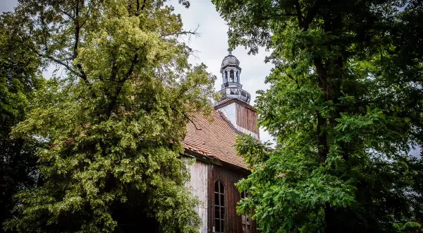 Stary kościół w Miejskiej Górce, otoczony bujną zielenią drzew, z charakterystyczną wieżą i drewnianą elewacją.