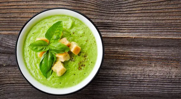 Un cremoso passato di verdure verde brillante, guarnito con crostini dorati e basilico fresco, su un tavolo di legno rustico.