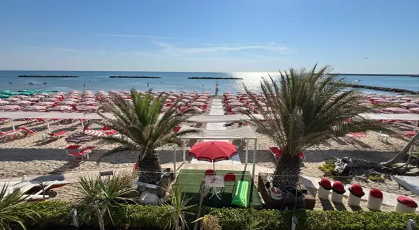 Spiaggia di Porto San Giorgio mare con file ordinate di ombrelloni rossi e bianchi, lettini, palme e il mare azzurro sotto un cielo limpido.