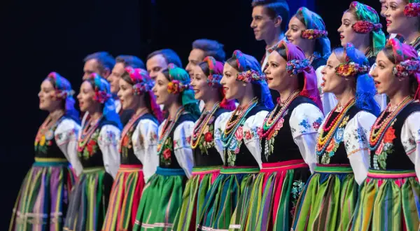 Zespół ludowy w tradycyjnych strojach, kobiety w kolorowych chustach i spódnicach, mężczyźni w tle.