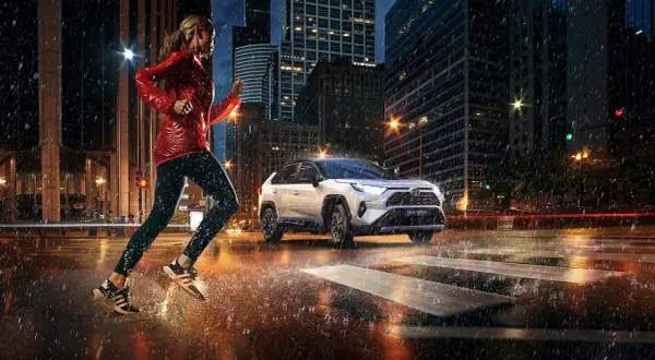 Kobieta biegnie w deszczu po mieście, obok niej jedzie biała Toyota RAV4 Hybrid. Zastanawiasz się, ile pali Toyota RAV4?