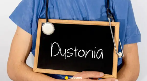 Lekarz w niebieskiej koszuli trzyma tabliczkę z napisem "Dystonia". Stetoskop zwisa na jego ramieniu.