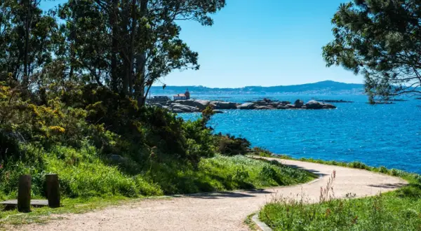 Sendero costero en la Illa de Arousa con mar azul brillante, rocas y un pequeño faro.