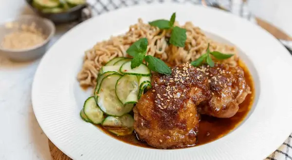 Två saftiga bitar sticky chicken med sesamfrön, serverade med nudlar och gurksallad.