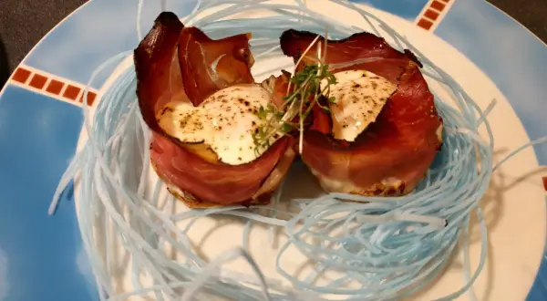 Zwei Eier mit Speck, kunstvoll in einem Nest aus blauen Fäden auf einem Teller angerichtet.