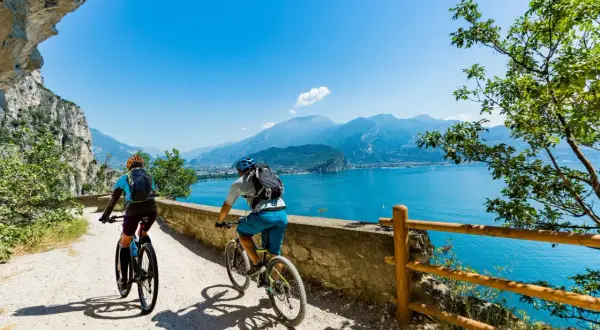 Ciclisti su una strada panoramica lungo il Lago di Garda, con montagne sullo sfondo.