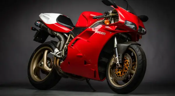 Czerwony motocykl Ducati z białym siedzeniem i złotymi felgami na ciemnym tle.
