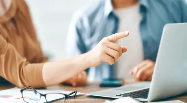 Dłoń wskazująca palcem na ekran laptopa, obok okulary i wykresy.