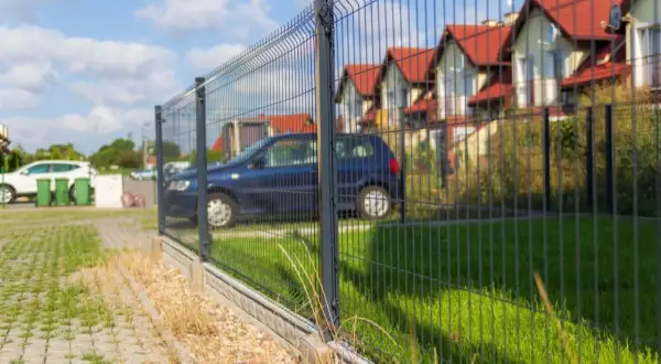 Płot z siatki z widocznym rozstawem słupków pod furtkę. W tle domy i samochody.