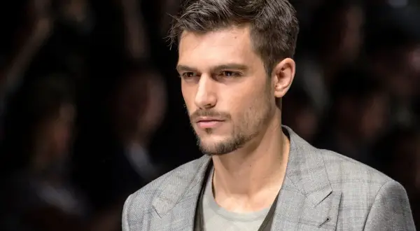 Uomo con capelli spettinati corti, sguardo intenso, indossa una giacca a quadri grigia.