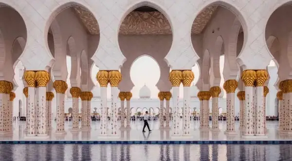 Un hombre camina bajo arcos blancos y dorados en Abu Dabi, reflejándose en el agua. Un visado Abu Dabi es el primer paso para disfrutar de esta maravilla.
