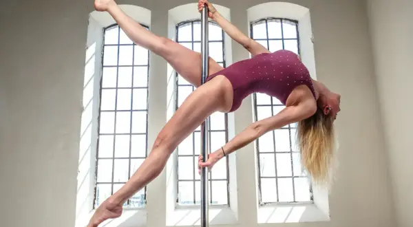 Kobieta w bordowym stroju wykonuje figurę na rurze pole dance, z nogami uniesionymi w powietrzu.