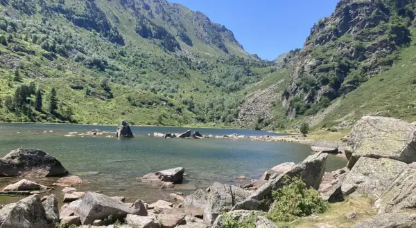 Lac de montagne entouré de montagnes verdoyantes et de rochers, sous un ciel bleu clair.