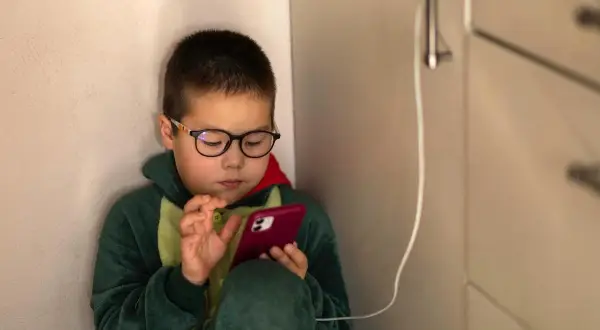 Chłopiec w okularach i piżamie z dinozaurem skupiony na telefonie. Czas na ekranie to wyzwanie, ale warto pamiętać o ograniczeniu czasu na telefonie.