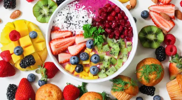 Kolorowa miska smoothie z owocami: truskawki, kiwi, mango, jagody, maliny, jeżyny, granat. Obok muffinki i migdały. Zdrowe produkty, które mogą zawierać histaminę.
