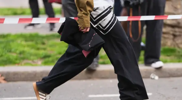Persona con un outfit casual, pantaloni neri ampi, giacca color kaki e scarpe da ginnastica Adidas marroni.