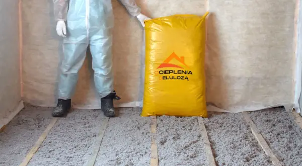 Osoba w kombinezonie ochronnym stoi obok worka z materiałem do ocieplenia celulozą. Podłoga pokryta jest warstwą izolacji.