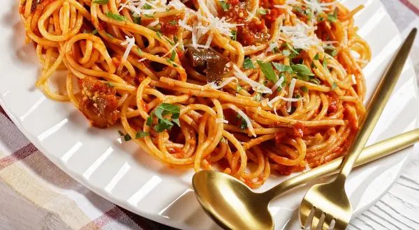 Spaghetti z bakłażanem i pomidorami, polane bogatym sosem sycylijskim, posypane parmezanem i natką pietruszki. Złote sztućce czekają.