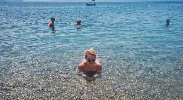 Kobieta w okularach przeciwsłonecznych leży w płytkiej wodzie na kamienistej plaży. W tle widać innych kąpiących się i łódź.